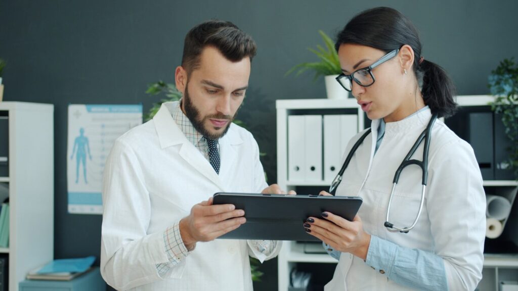 Two healthcare professionals, one male and one female, review information on a tablet in a modern office setting, indicating collaboration in patient care.