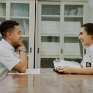 Two students, a young man and woman, are engaged in a lively conversation at a table in a library, sharing a book between them.