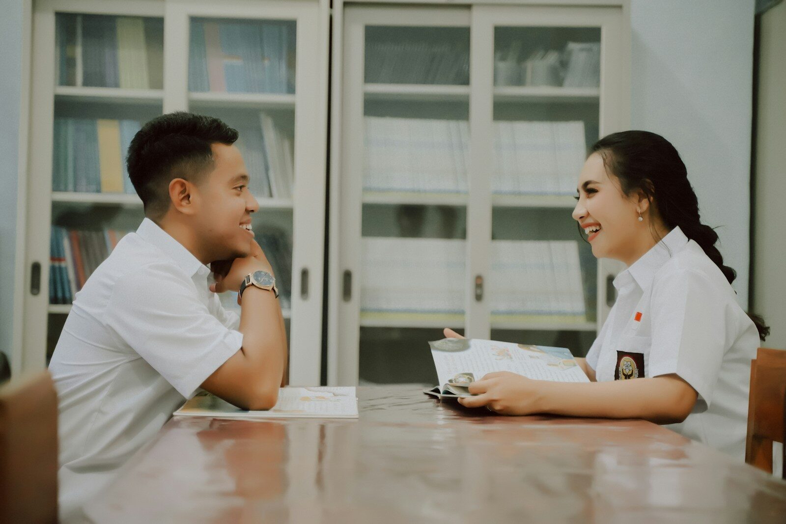 Two students, a young man and woman, are engaged in a lively conversation at a table in a library, sharing a book between them.