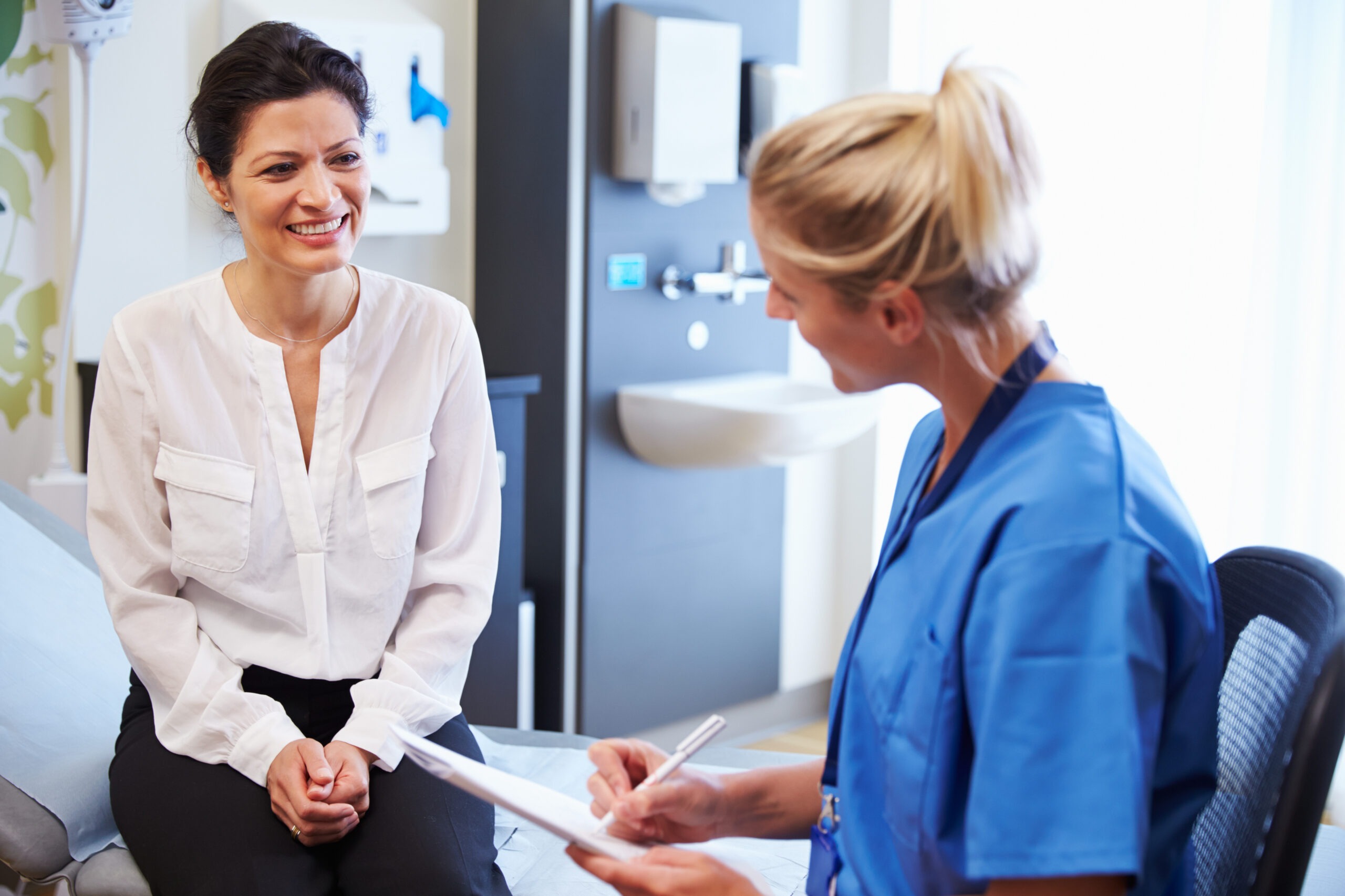 A healthcare professional in blue scrubs discusses medical notes with a smiling patient in a clinical setting, indicating a routine check-up.